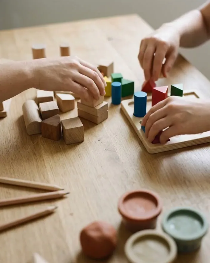 Hände bauen mit Holzklötzen und Formen an einem Holztisch, davor Knete und Stifte
